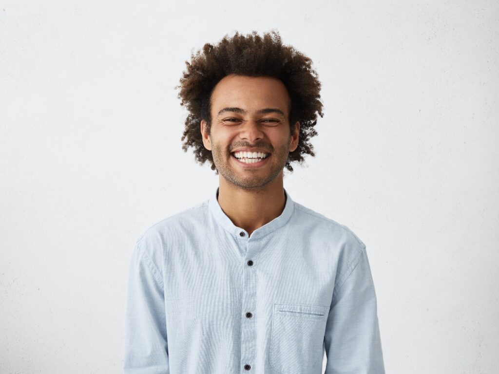 Handsome unshaven young dark-skinned male laughing out loud at funny meme he found on internet, smiling broadly, showing his white straight teeth. Positive human facial expressions and emotions