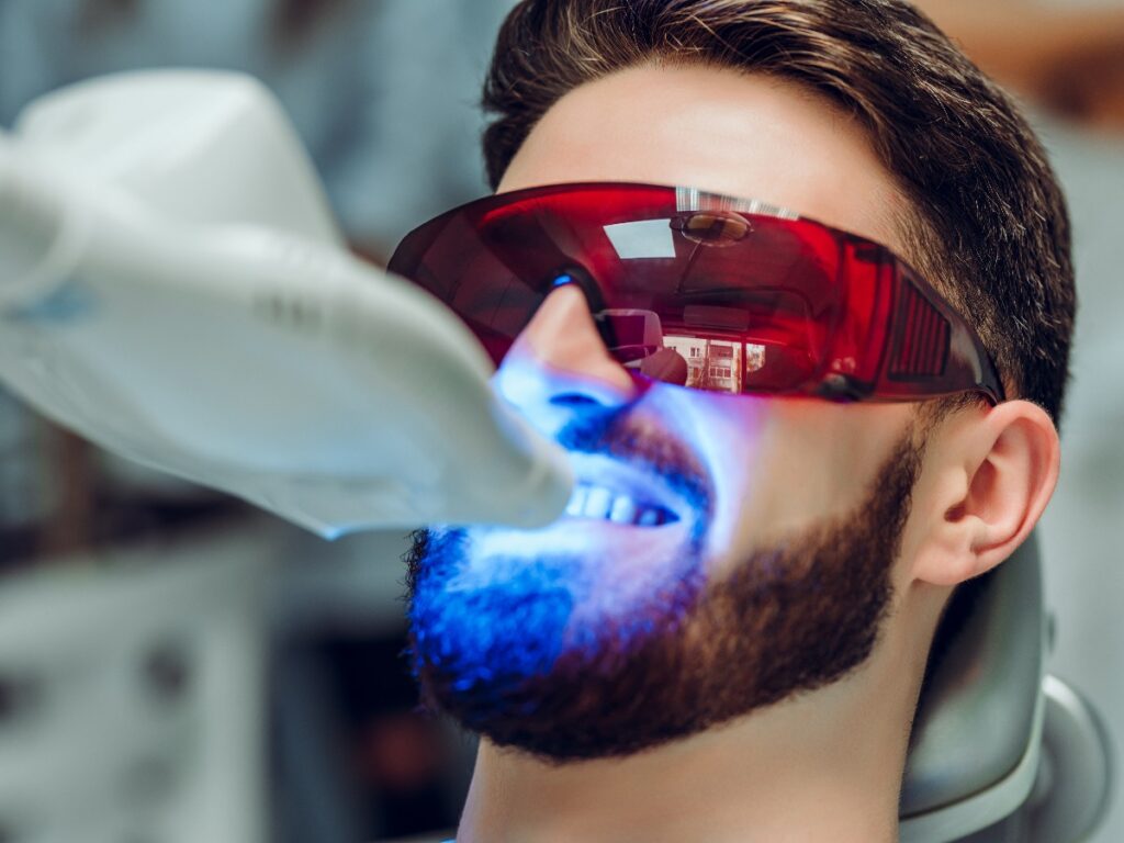Man having teeth whitening by dental UV whitening device,dental assistant taking care of patient,eyes protected with glasses. Whitening treatment with light, laser, fluoride.