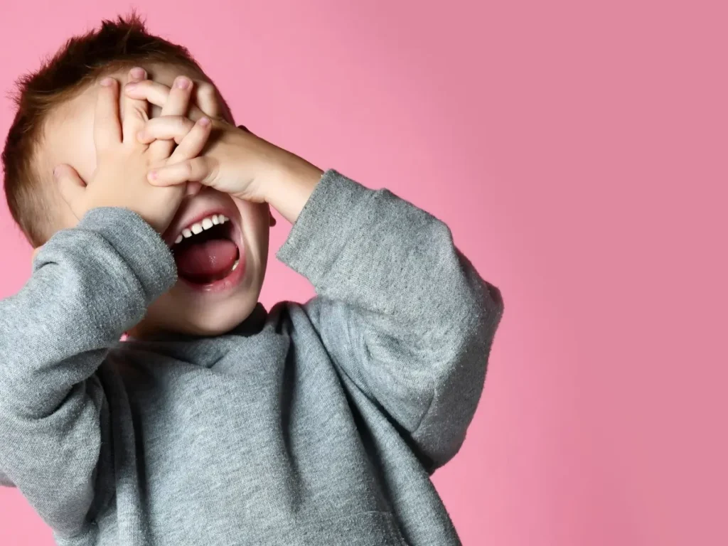 Ein fröhlicher Junge in grauem Pullover lacht ausgelassen und bedeckt spielerisch sein Gesicht mit den Händen vor rosa Hintergrund